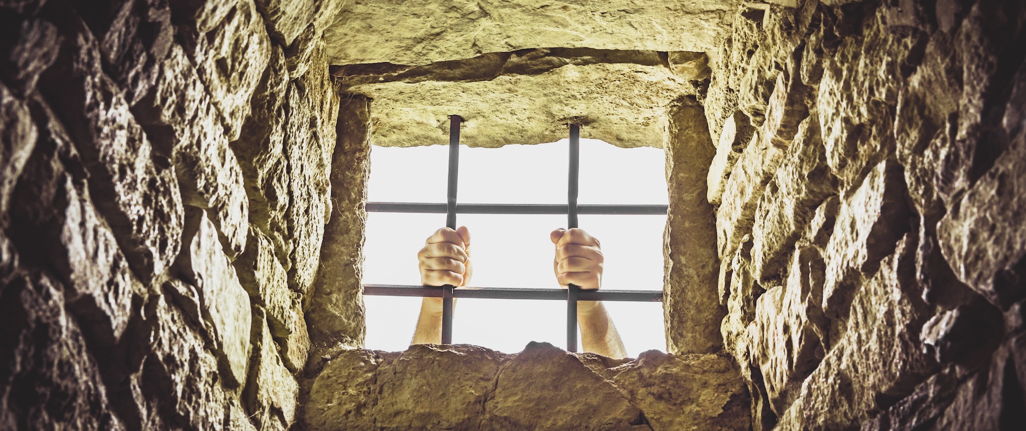A pair of hands holding bars at the window of a stone jail