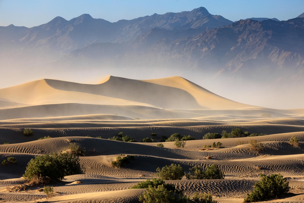 A landscape including mountains in the background, sand dunes in the middle ground and bushes in the foreground